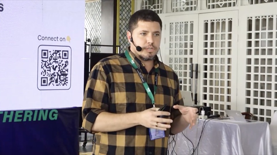 Isaiah on stage mid-sentence, headset mic on, one hand open toward the audience, a white latticed door behind him.